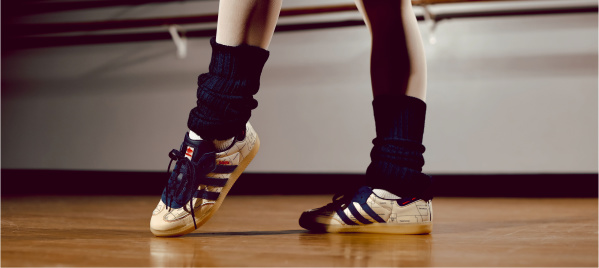 Close-up of a dancer’s legs and feet in sneakers and leg warmers, poised on a wooden studio floor near a ballet barre.