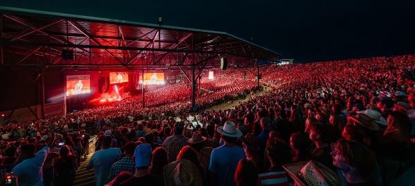 An open-air concert stage with thousands of people watching a show illuminated in a red glow from the stage lights against the pitch black of the night sky.
