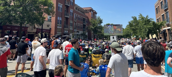 A large crowd gathers outside Three Lions Pub on a sunny day to watch a sporting event on a giant outdoor screen set up in the middle of the street. Fans wearing jerseys, hats and team gear fill the block, with many seated in folding chairs while others stand shoulder to shoulder. Brick apartment buildings and storefronts frame the scene, and the atmosphere resembles a lively neighborhood watch party.