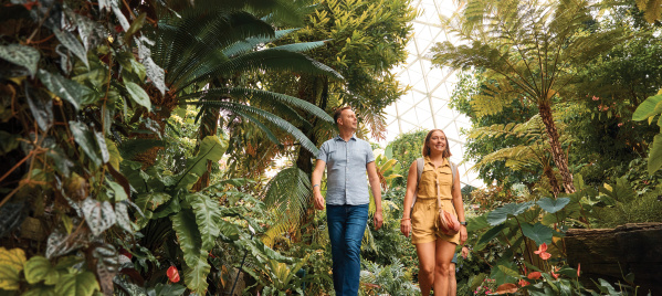 Two adults walk along a lush indoor garden path beneath a glass-domed ceiling, surrounded by tall palms, ferns and tropical plants inside a conservatory.