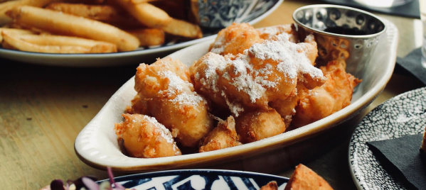 A cozy brunch spread featuring crispy potatoes, eggs Benedict, powdered sugar–dusted fritters with dipping sauce, and a sandwich with fries, all paired with iced tea and water on a sunlit table.