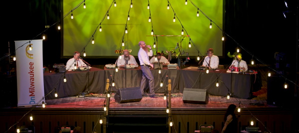 A lively stage event with a panel of speakers seated at tables under hanging string lights, facing a large audience in a dimly lit venue with a bright green backdrop screen.