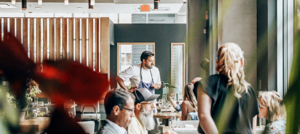 Diners enjoy a meal inside a bright, modern Milwaukee restaurant with large windows, warm lighting, and a relaxed, welcoming atmosphere as a chef engages with guests.