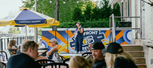A woman in a navy blue romper singing into a microphone on a stage, holding a pink guitar, with people in front of her sitting at tables under gold and blue umbrellas on a patio along the riverfront with trees in the background.