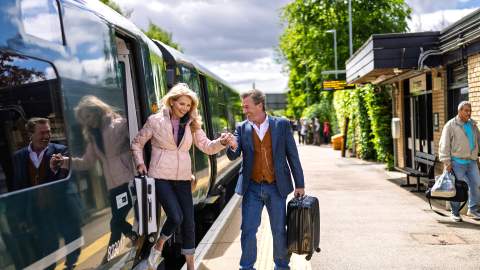 A couple disembark a train carrying suitcases at Kingham station