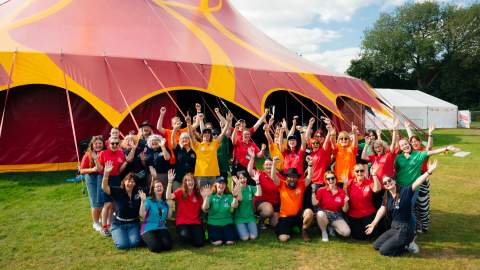 The team from Big Malarkey Festival in colourful t shirts in a group in front of a big red and yellow marquee.