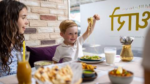 Child holding up noodles on a fork around table with family at Padharo