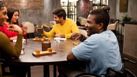 A group of people sit eating, drinking and laughing at a table in a cafe. One of the people is using a wheelchair.