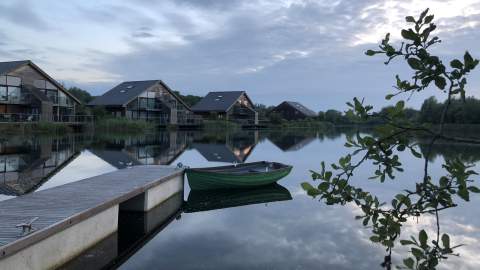 A green rowing boat is tied to a jetty on a lake, in the distance are modern villas with large windows positioned on the waterfront, at Cotswold Water Park Retreats in the Cotswolds