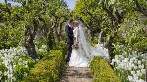 A bride and groom embrace under an archway of trees with white tulip borders at Lords of the Manor