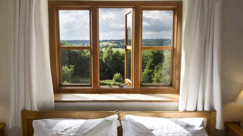 A bed with white linen sits in front of a window framed by crisp white curtains. Through the window you see trees and rolling hills under a cloudy sky. At Heath Farm Holiday Cottages in the Cotswolds