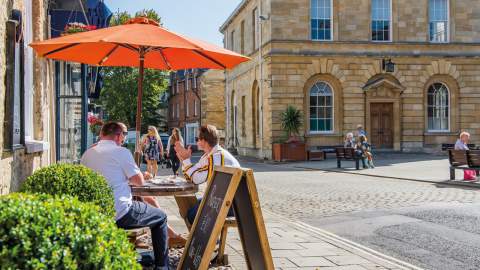 A group of people sit in the shade of an orange parasol outside a cafe, people sit on benches outside the adjacent town hall, whilst two ladies walk along shopping. Woodstock in the Cotswolds
