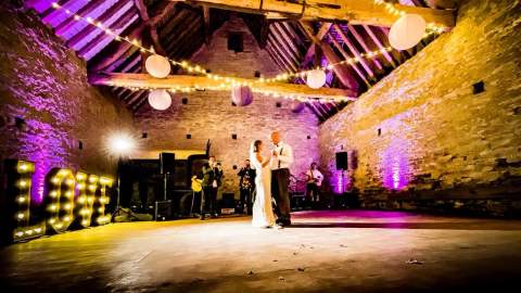 A newly married couple dance in the centre of a historic barn with fairy lights hanging from the wooden beamed ceiling at Cogges Manor Farm in the Cotswolds