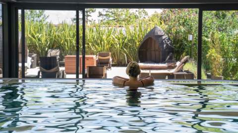 A person relaxes in an indoor pool, facing large glass doors that reveal a lush garden with wicker furniture and greenery, creating a tranquil atmosphere.
