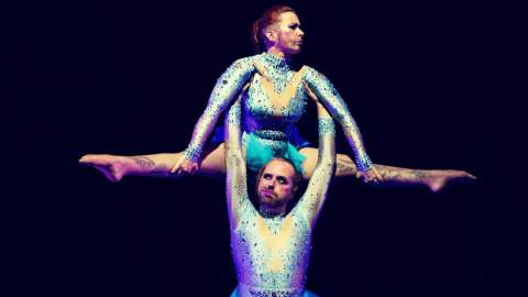 Two people wearing blue leotards in an acrobatic position. One person is holding another person in their air under their armpits.