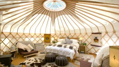 A circular skylight illuminates a cozy double bed, faux animal skin rugs and pouffes dotted in front of a wood burner inside Sapperton Yurt, at Westley Farm in the Cotswolds