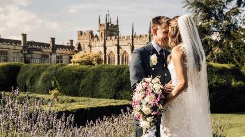 A bride and groom stand face-to-face, smiling, in a garden with Sudeley Castle in the background