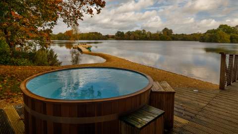 A wooden hot tub on a deck with views across the lake and leaves falling from the trees, at Log House Holidays in the Cotswolds