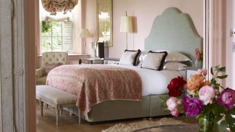 A vase of pink peonies sits on a table in front of a view of a large bedroom with upholstered bed, pale pink walls and large sash window at Thyme in the Cotswolds