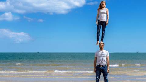 Two people on a beach wearing blue jeans and white tops. One person is standing on the head of another person.