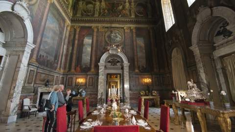 A couple with a tour guide stand in an impressive room with painted images on the ceiling and walls, large marble doorways and gold furniture at Blenheim Palace