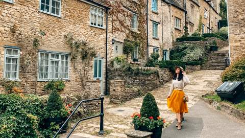 A woman wearing a hat smiles as she poses in front of stone cottages on a stepped hill in Tetbury
