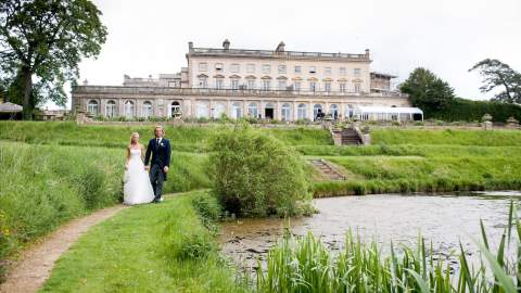 A bride and groom walk beside a lake with Cowley Manor in the background