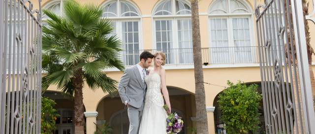 A couple in wedding attire shares a romantic moment amidst vibrant greenery and a charming building in the background.