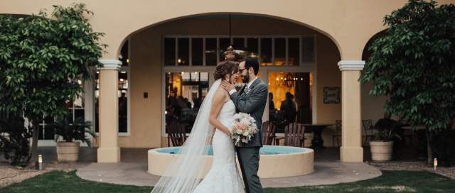 Groom in gray tuxedo kisses bride in mermaid-style wedding dress with veil on the forehead standing on the grass of the Crowne Plaza courtyard. A fountain can be seen in the background along with orange trees, and the historic, tan Crowne Plaza building with large archways.