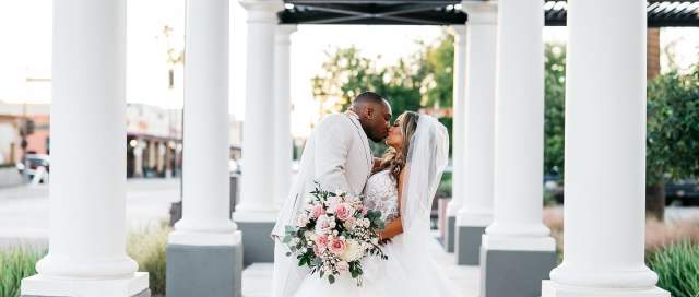 A newlywed couple shares a romantic kiss under a covered walkway, surrounded by white columns and lush greenery. The bride holds a bouquet of pink roses.