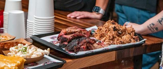 Pulled pork and brisket pilled on a platter on a wooden counter, with a waiter in the background, and sides on a platter off to the left.