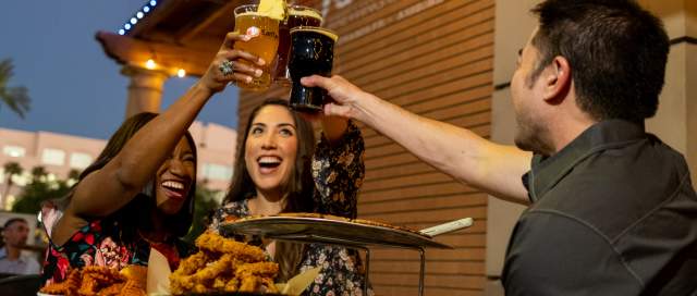 Two women and one man cheers beers on SanTan Brewing patio with food.