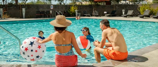 A family enjoying pool time at their hotel, with mom and dad in the foreground, backs to the camera, seated at the pool with their feet hanging over the edge into the pool, and their two young, smiling children facing them with pool toys.