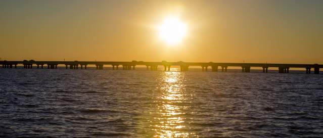 The Lake Pontchartrain Causeway Bridge stretches across Lake Pontchartrain at sunset, silhouetted against a golden sky. The low sun hangs above the bridge, casting a bright reflection across the rippling water as vehicles travel along the span between the Northshore and the Southshore.