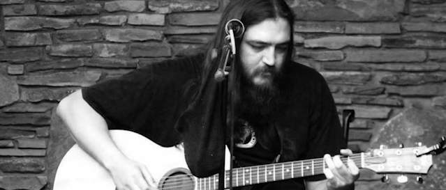 Black and white photo of a long-haired man sitting on a chair strumming a guitar