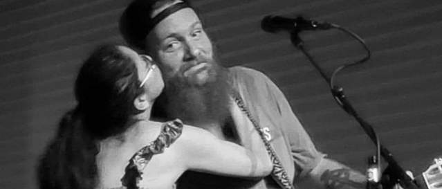 Black and white photo of a bearded man holding a guitar being kissed on the cheek by a woman in a long dress.