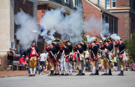 A group of historical reenactors in colonial military uniforms fires muskets, creating clouds of smoke during a lively outdoor event.