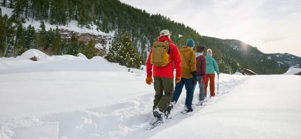Cross country skiiers trekking along a snowy path with forest covered mountains in the background.