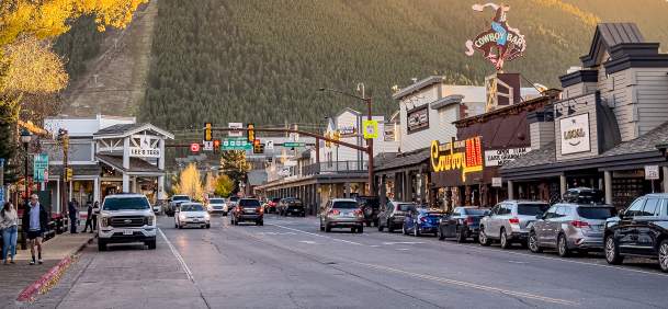 A western-style downtown street lined with shops and parked cars sits beneath forested mountains, with a large “Cowboy Bar” sign visible above the buildings in warm evening light.