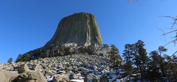 Devils Tower rises above a rocky, snow-covered landscape in northeastern Wyoming. Patches of winter snow cover the boulder field and surrounding pine forest beneath the monument’s distinctive vertical rock columns and a clear blue sky.