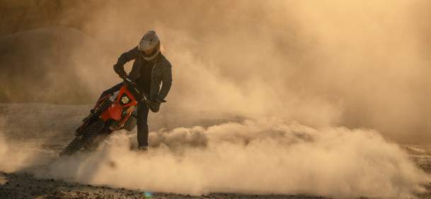 A motorcyclist rides across a dusty dirt road, kicking up a cloud of dust against a warm, sunlit landscape.