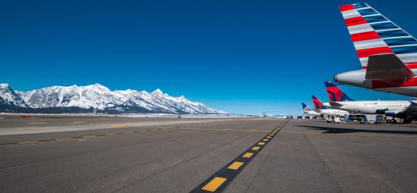Airplanes lined up on a runway with snow-capped mountains in the background under a clear blue sky. The scene captures a vibrant travel hub in Jackson Wyoming.