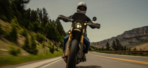 A motorcyclist rides along a winding mountain road, with forested hills and rocky cliffs in the background under a clear sky.