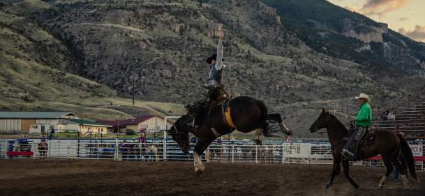 A rodeo rider raises one arm while riding a bucking horse inside an outdoor arena, with another mounted rider nearby and tree-covered mountains rising in the background at sunset.
