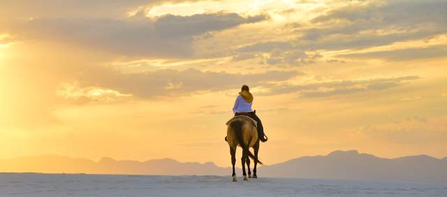a person rides a horse through white sand dunes before a sky painted with the orange light of a setting sun