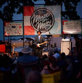 Booker T. Jones stands on the stage with an electric guitar. The crowd is seen from behind in the foreground. The backdrop shows a list of sponsor logos and in the center is the Music on Main 2026 logo.
