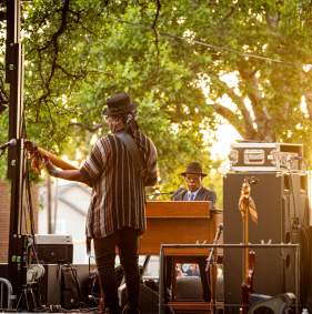 Booker T. Jones (on the piano) and his band members. The image is taken from the back of the stage and there is music equipment and speakers throughout the stage.