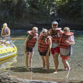 Four rafters wearing lifejackets stand ankle deep in the shallow river water beside a yellow raft with their guide.