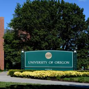 Large Green University of Oregon Campus Sign with gold seal in front of red brick building.