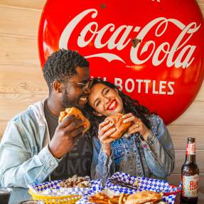 Couple eating burgers with a Coca-Cola sign behind them